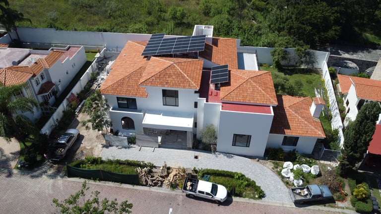 Vista aérea de una casa moderna con techo de teja naranja y paneles solares, rodeada de jardinería y autos estacionados