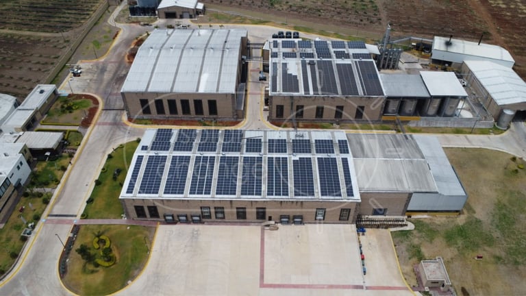 Aerial view of industrial buildings with solar panels on roofs and parking area surrounded by green landscaping.